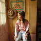 A young girl with a braid sits on a stool by a window, wearing the Louise Misha Girls’ Acacia Sweater—featuring handmade embroidery—and striped pants in a cozy, vintage-inspired room with hats hanging on the wall.
