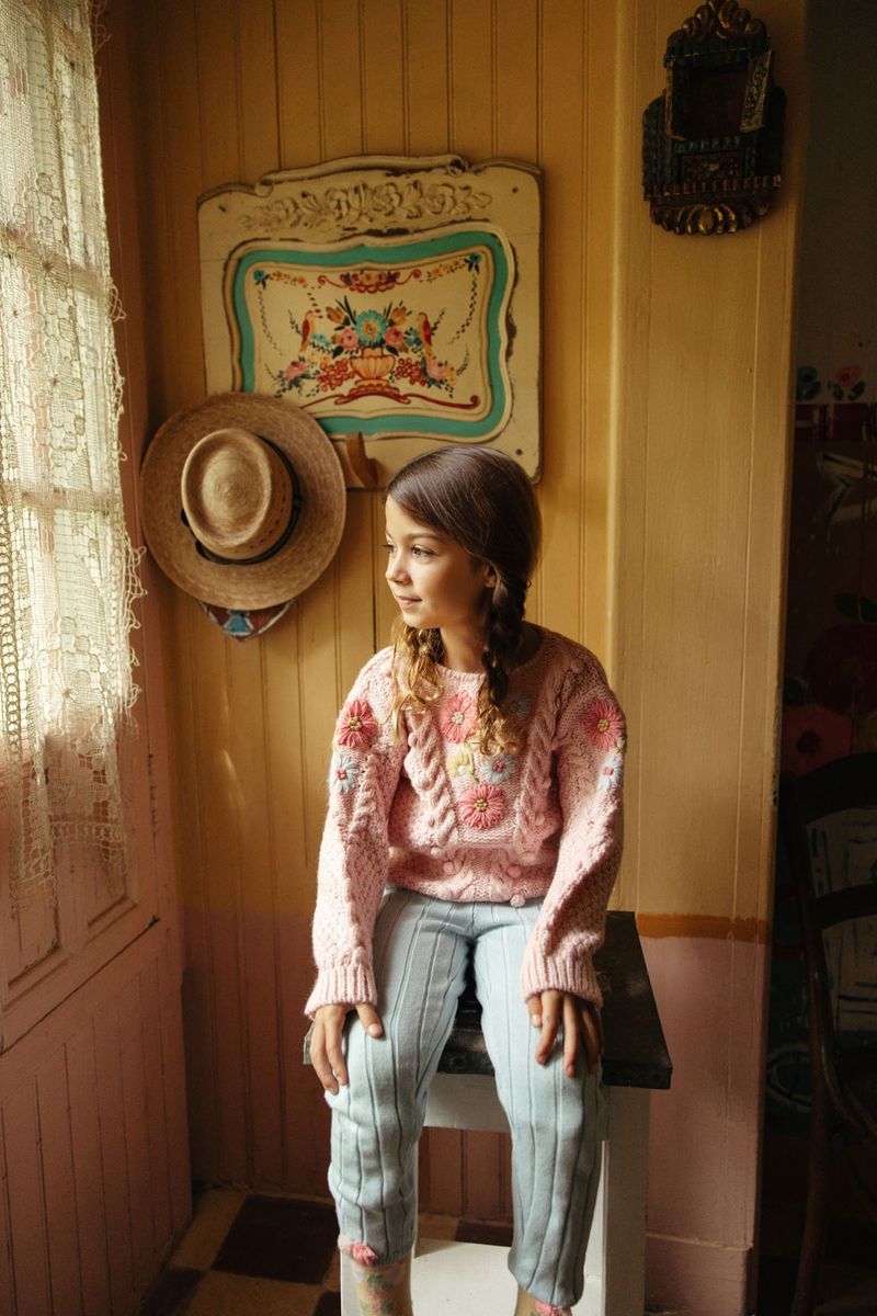 A young girl with a braid sits on a stool by a window, wearing the Louise Misha Girls’ Acacia Sweater—featuring handmade embroidery—and striped pants in a cozy, vintage-inspired room with hats hanging on the wall.