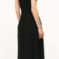 A person with wavy, chin-length hair stands with their back to the camera, wearing Staud's sleeveless black Elyse Dress with a crew neckline, pleated skirt, and cream knee-high boots.