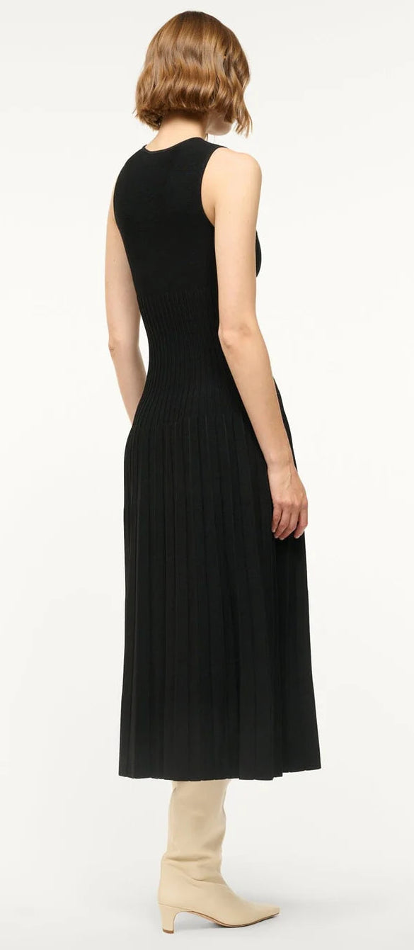 A person with wavy, chin-length hair stands with their back to the camera, wearing Staud's sleeveless black Elyse Dress with a crew neckline, pleated skirt, and cream knee-high boots.