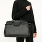 A woman wearing a black sweater and pants holds the Staud Maude Carryall, a large oversized bag crafted from pebbled leather with top handles and organizational pockets, set against a plain background.