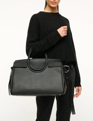 A woman wearing a black sweater and pants holds the Staud Maude Carryall, a large oversized bag crafted from pebbled leather with top handles and organizational pockets, set against a plain background.