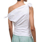A person is shown from the back against a white background, wearing the Staud Bellaria Top—a white one-shoulder evening top with tied details and an asymmetrical neckline—paired with light green shorts.