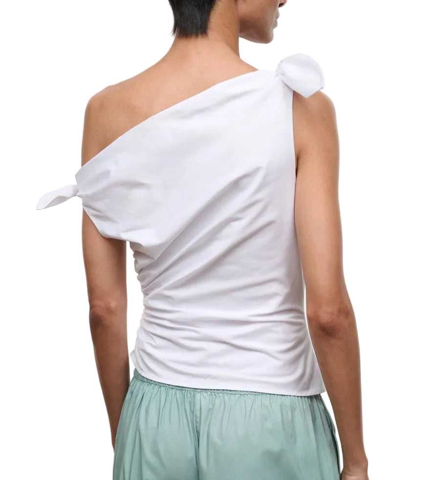A person is shown from the back against a white background, wearing the Staud Bellaria Top—a white one-shoulder evening top with tied details and an asymmetrical neckline—paired with light green shorts.