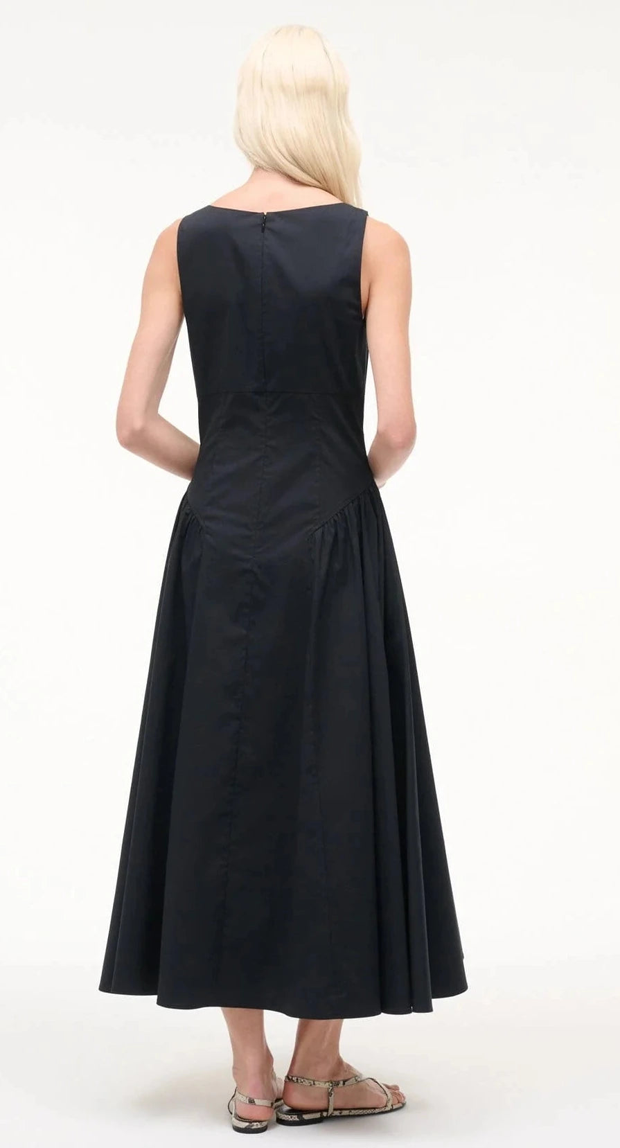 A person with long blonde hair stands with their back to the camera against a white background, wearing Staud’s sleeveless, ankle-length Gwen Dress in black with structured pleats and flat sandals.