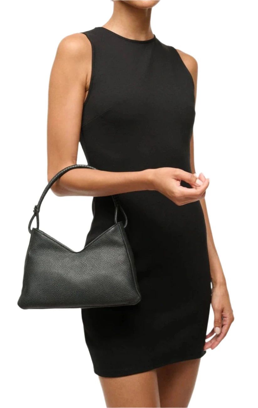 A person in a sleeveless black dress is holding the Staud Valerie Shoulder Bag, a sleek black pebbled leather accessory with a discreet crossbody strap.