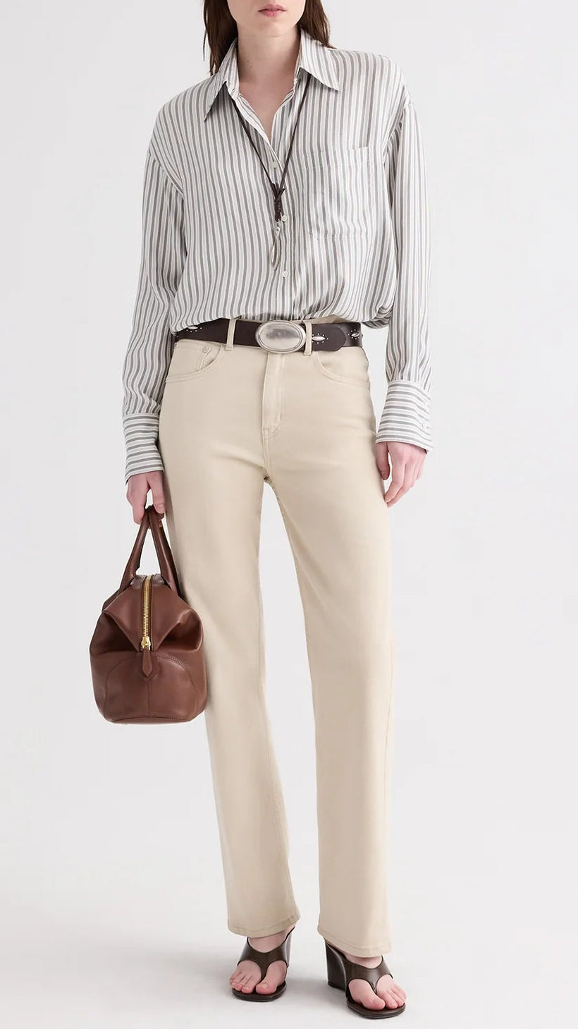 A person dressed in a striped shirt, beige pants, dark belt, and sandals holds the Small Howard Bag by TWP in Cognac leather, lending a refined touch and structured silhouette to the ensemble.