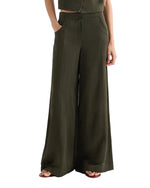 Model wears TWP's Demie Coated Viscose Linen Pant in Rosen Green, a high-waisted, wide-leg style, with hands in pockets and sandals. Photo shows shoulders to feet against a white background.