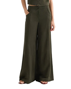 Model wears TWP's Demie Coated Viscose Linen Pant in Rosen Green, a high-waisted, wide-leg style, with hands in pockets and sandals. Photo shows shoulders to feet against a white background.