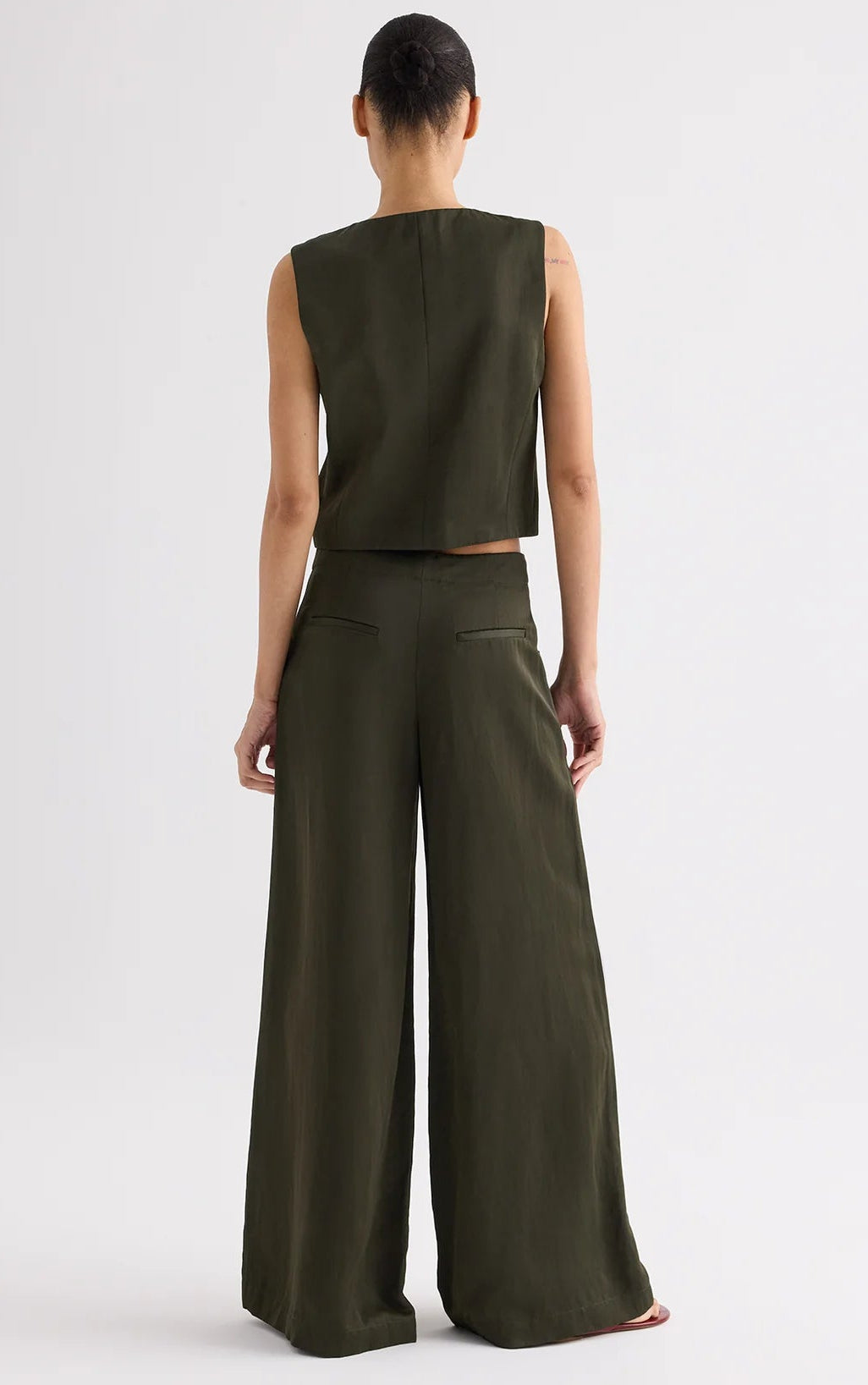 A person faces away from the camera, wearing a sleeveless dark top and matching TWP Demie Coated Viscose Linen Pant with a wide-leg silhouette.