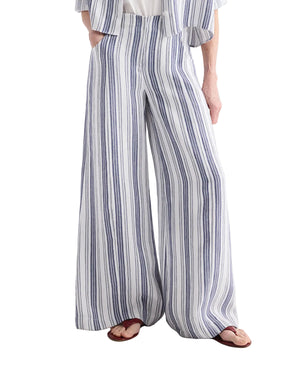 Person wearing the TWP Demie Linen Stripe Pant, featuring a blue and white vertical stripe pattern and wide-leg, high-waisted silhouette, paired with brown sandals. Only the lower half of the body is visible.