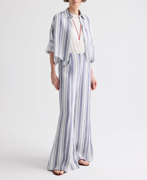 A mature woman stands against a plain background wearing the TWP Demie Linen Stripe Pant, featuring a blue and white vertical stripe, paired with a matching shirt, a simple white top, a red necklace, and sandals.