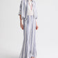 A person wearing the TWP Next Ex Linen Stripe Shirt in Ivory/Navy—a loose-fitting, blue-and-white vertical striped piece—layered over a white top with long pants, accessorized with a pendant necklace and sandals, stands against a plain white background.