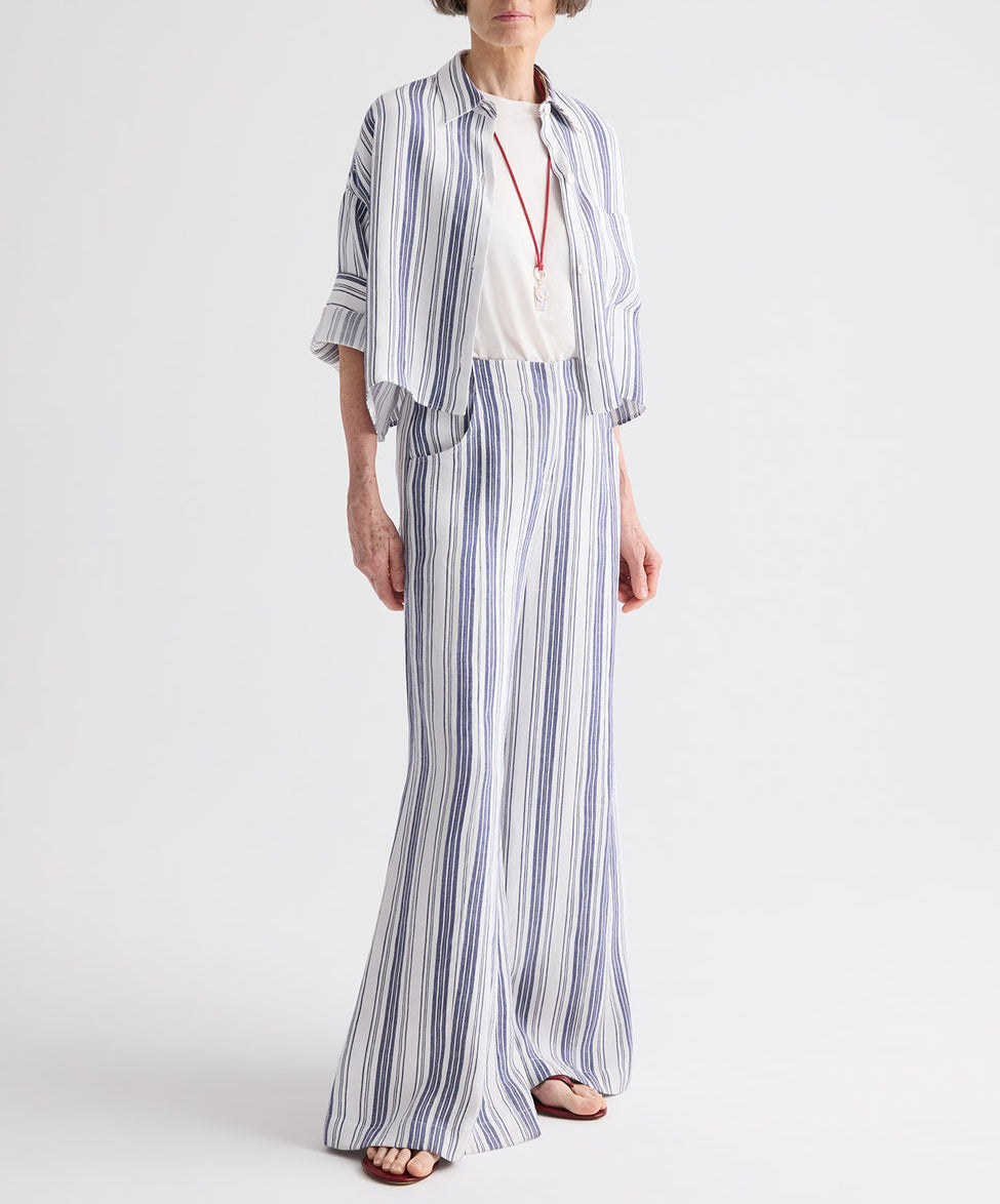 A person wearing the TWP Next Ex Linen Stripe Shirt in Ivory/Navy—a loose-fitting, blue-and-white vertical striped piece—layered over a white top with long pants, accessorized with a pendant necklace and sandals, stands against a plain white background.