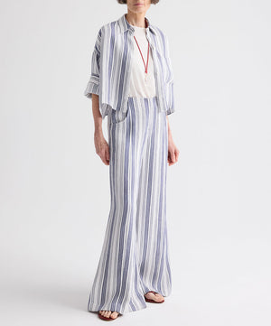 A person wearing the TWP Next Ex Linen Stripe Shirt in Ivory/Navy—a loose-fitting, blue-and-white vertical striped piece—layered over a white top with long pants, accessorized with a pendant necklace and sandals, stands against a plain white background.