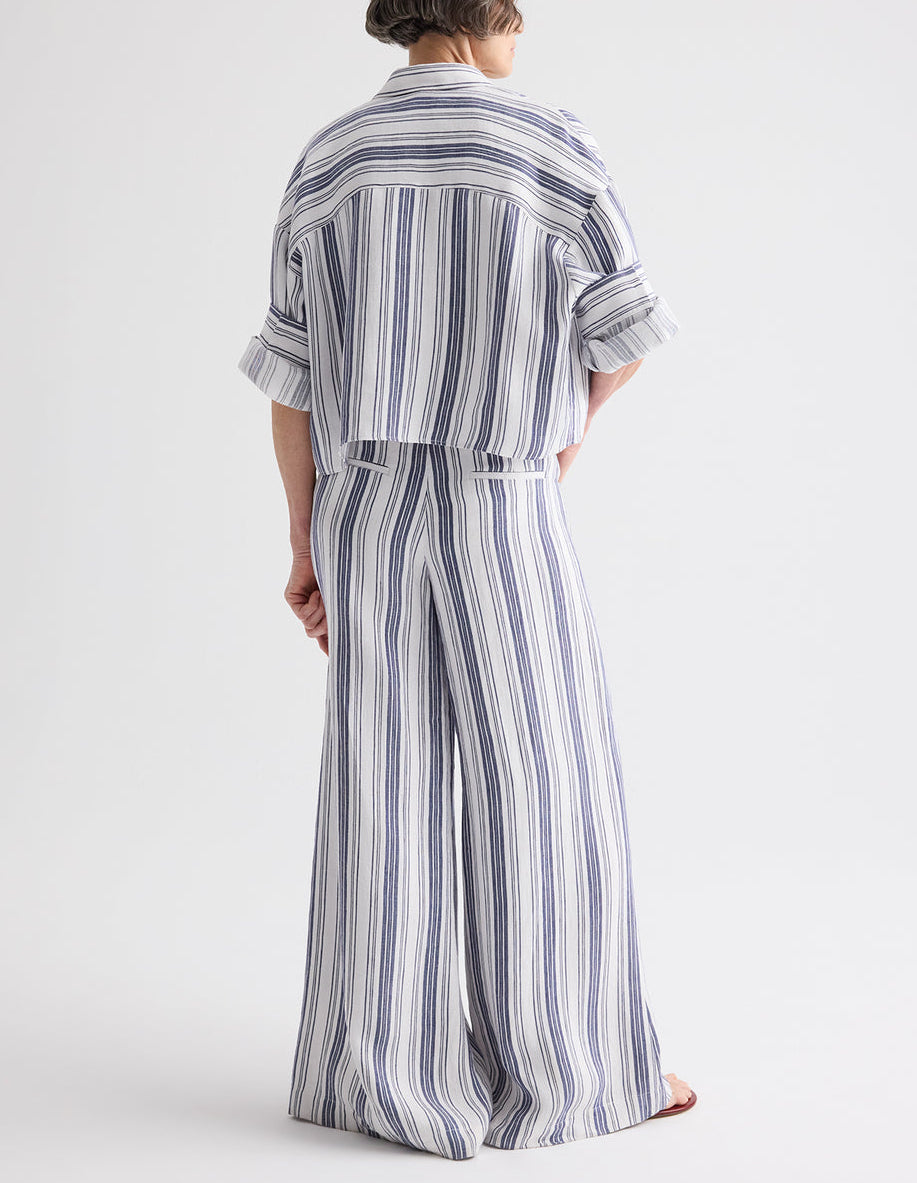 A person stands with their back to the camera, wearing a loose-fitting blue and white vertical stripe shirt paired with TWP's Demie Linen Stripe Pant.