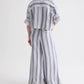 A person stands with their back to the camera, wearing loose-fitting blue and white vertically striped pants and the TWP Next Ex Linen Stripe Shirt—a matching ivory/navy cropped short-sleeve button-down shirt with rolled sleeves.