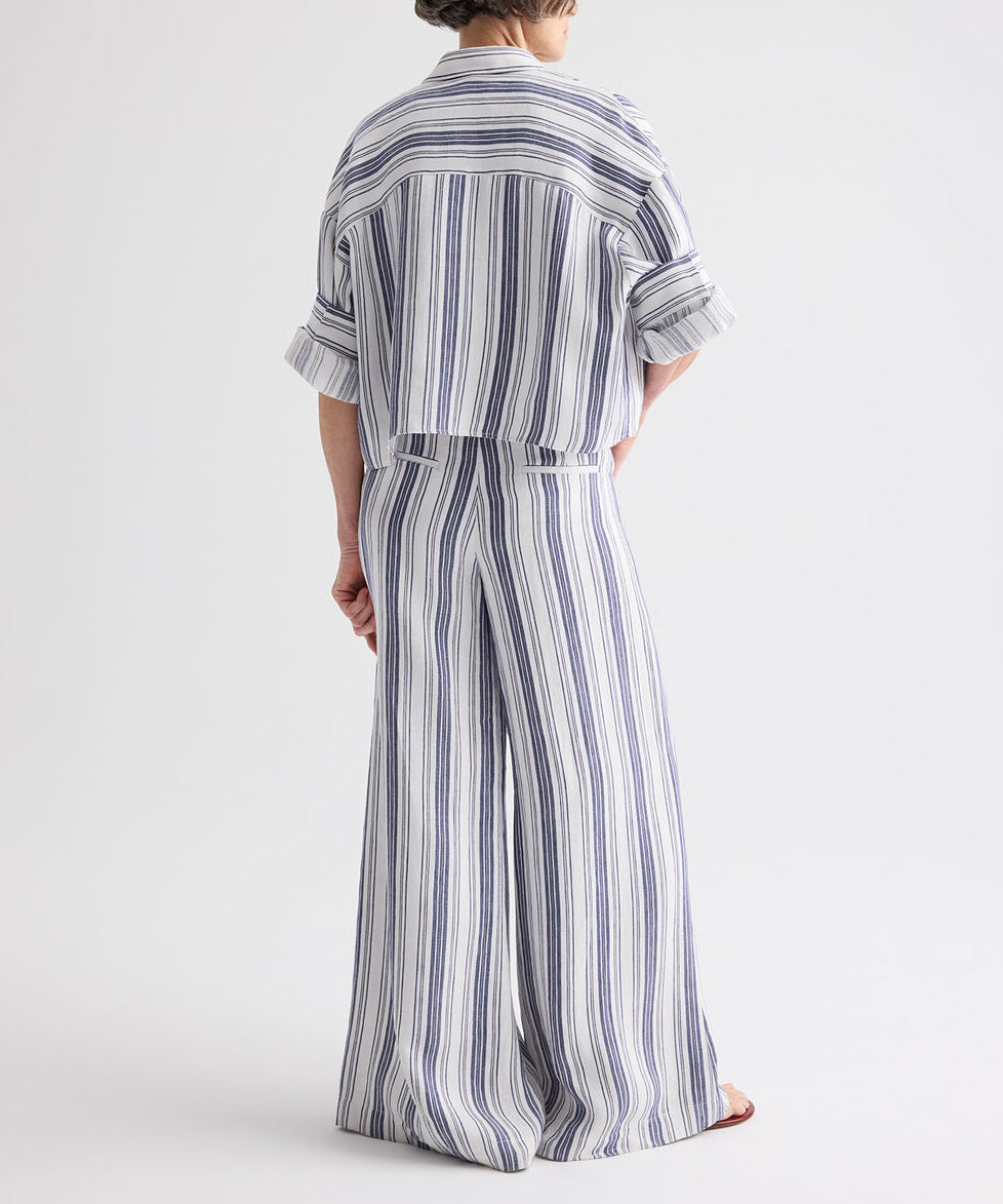 A person stands with their back to the camera, wearing loose-fitting blue and white vertically striped pants and the TWP Next Ex Linen Stripe Shirt—a matching ivory/navy cropped short-sleeve button-down shirt with rolled sleeves.