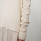 A person wears the Lisa Yang Mila Cashmere Sweater, a loose, long-sleeved cream knit, paired with a light beige relaxed skirt, standing against a plain background.