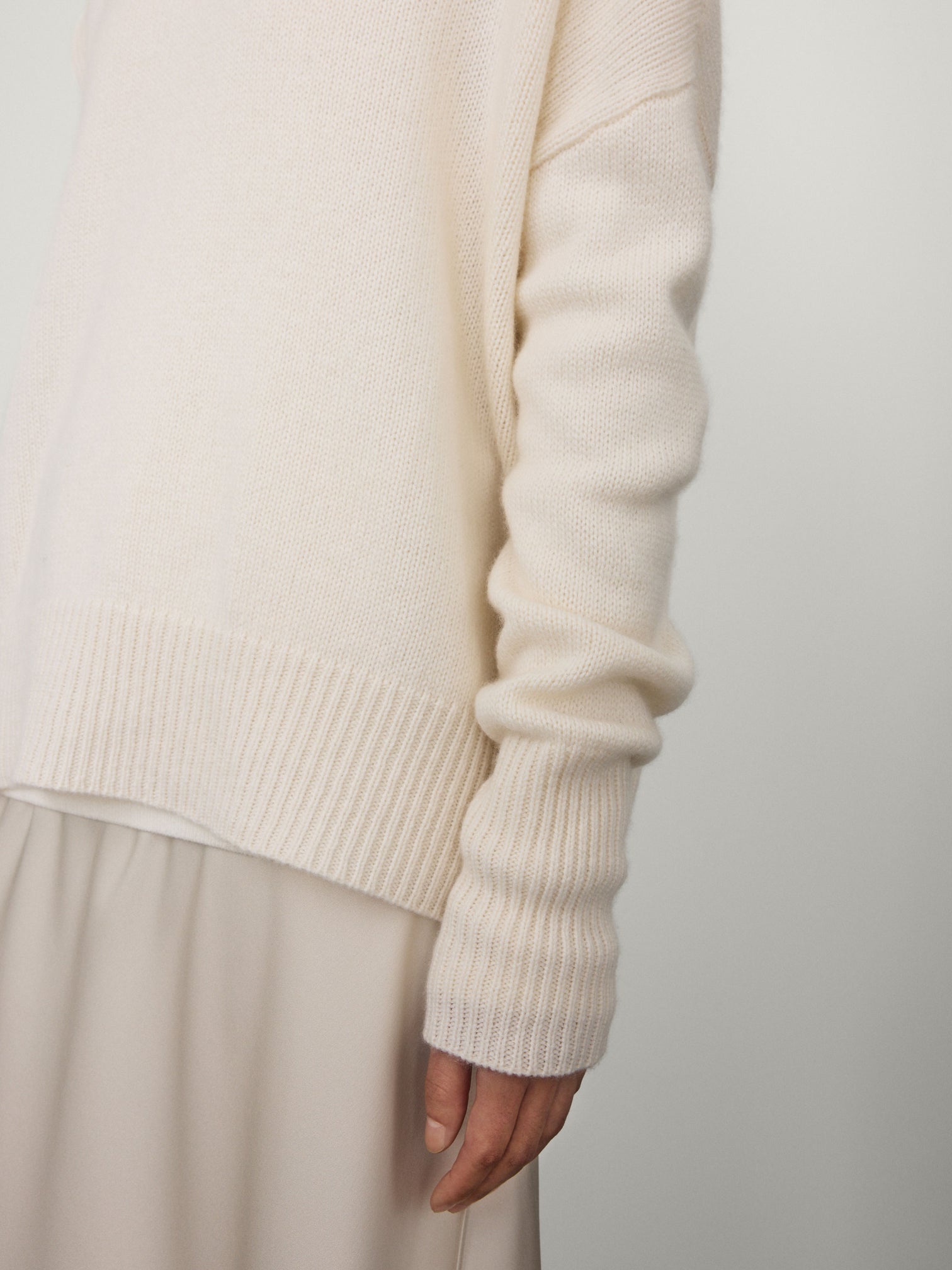 A person wears the Lisa Yang Mila Cashmere Sweater, a loose, long-sleeved cream knit, paired with a light beige relaxed skirt, standing against a plain background.
