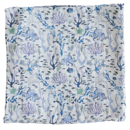 The Timo & Violet Shawl Blanket is a square cloth with a blue and purple underwater coral reef pattern, featuring fish, seaweed, octopuses, and marine motifs on a light background—ideal as a baby accessory.