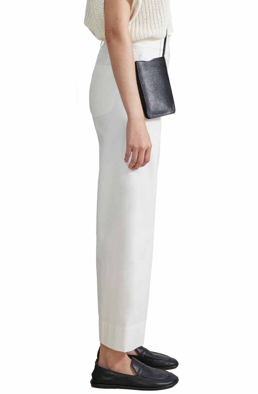 Person standing in profile view, wearing a white knit top, Apiece Apart Classic Merida Pant by Apiece Apart, black loafers, and a small black crossbody bag.