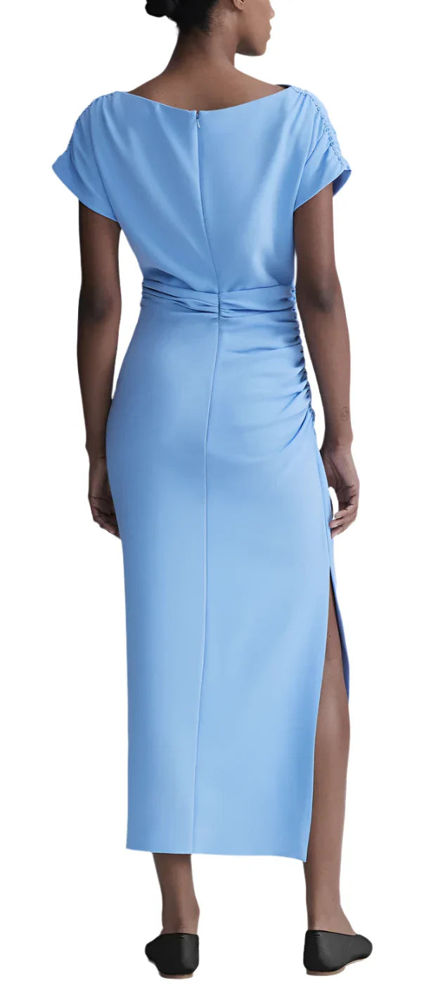 A woman wears the Lela Rose Florence Side Ruched Fluid Crepe Dress, featuring a floor-length blue design with elegant goddess ruching and short sleeves, paired with black shoes, viewed from behind.