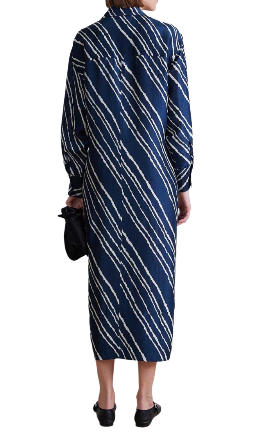 Adorned in the Apiece Apart Mila Silk Shirt Dress, an elegantly styled person showcases timeless sophistication. This long blue silk dress, embellished with white diagonal stripes and a self-tie waist, perfectly complements the black clutch they hold while facing away.