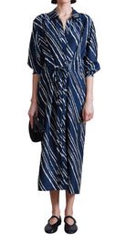 A person is wearing the Apiece Apart Mila Silk Shirt Dress, a long-sleeved blue and white striped silk dress with a button closure and self-tie waist, accessorized with a small black clutch and matching black shoes.