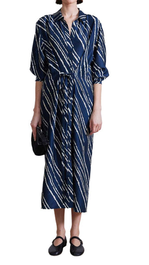 A person is wearing the Apiece Apart Mila Silk Shirt Dress, a long-sleeved blue and white striped silk dress with a button closure and self-tie waist, accessorized with a small black clutch and matching black shoes.