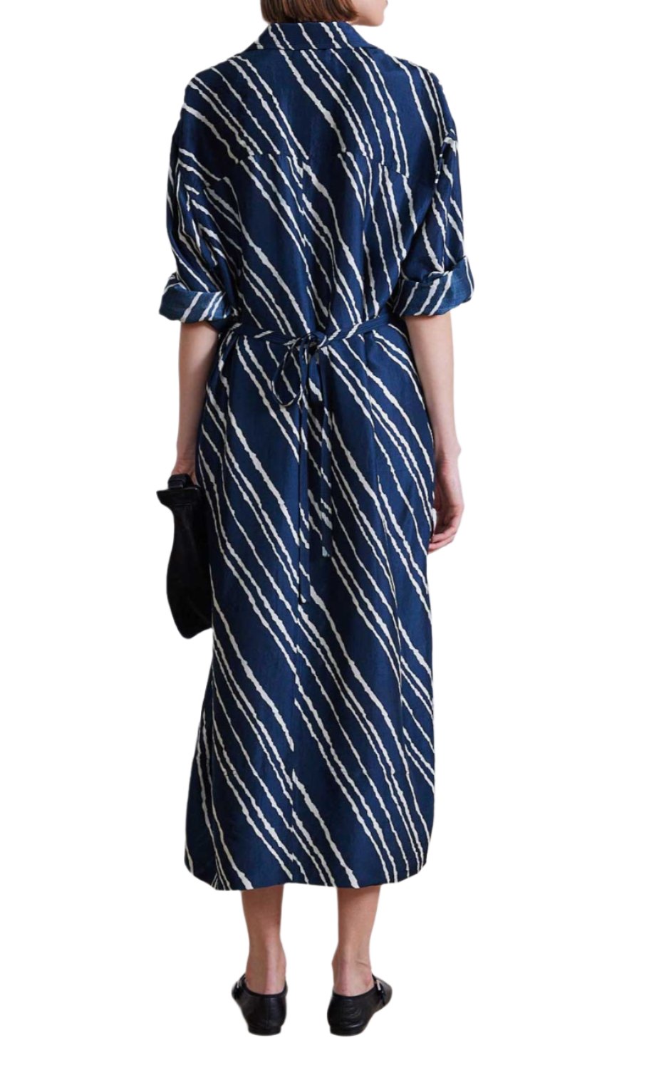 A person wearing the Apiece Apart Mila Silk Shirt Dress, which features a long blue and white patterned design with rolled sleeves and a self-tie waist, carries a black clutch. She completes her outfit with black shoes.