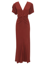 The Victoria Beckham Gathered V Neck Midi Dress by Victoria Beckham in rust-colored viscose crepe fabrication features short puff sleeves, a waist-defining pleat detail with a twisted knot at the waist, and a knee-high slit at the front.