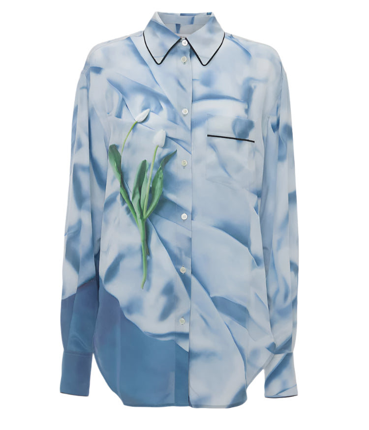 The Victoria Beckham Piping Detail Pyjama Shirt is a long-sleeve silk shirt with a blue and white abstract floral print, collar, and black-trimmed pocket, featuring an elegant tulip design on the side.