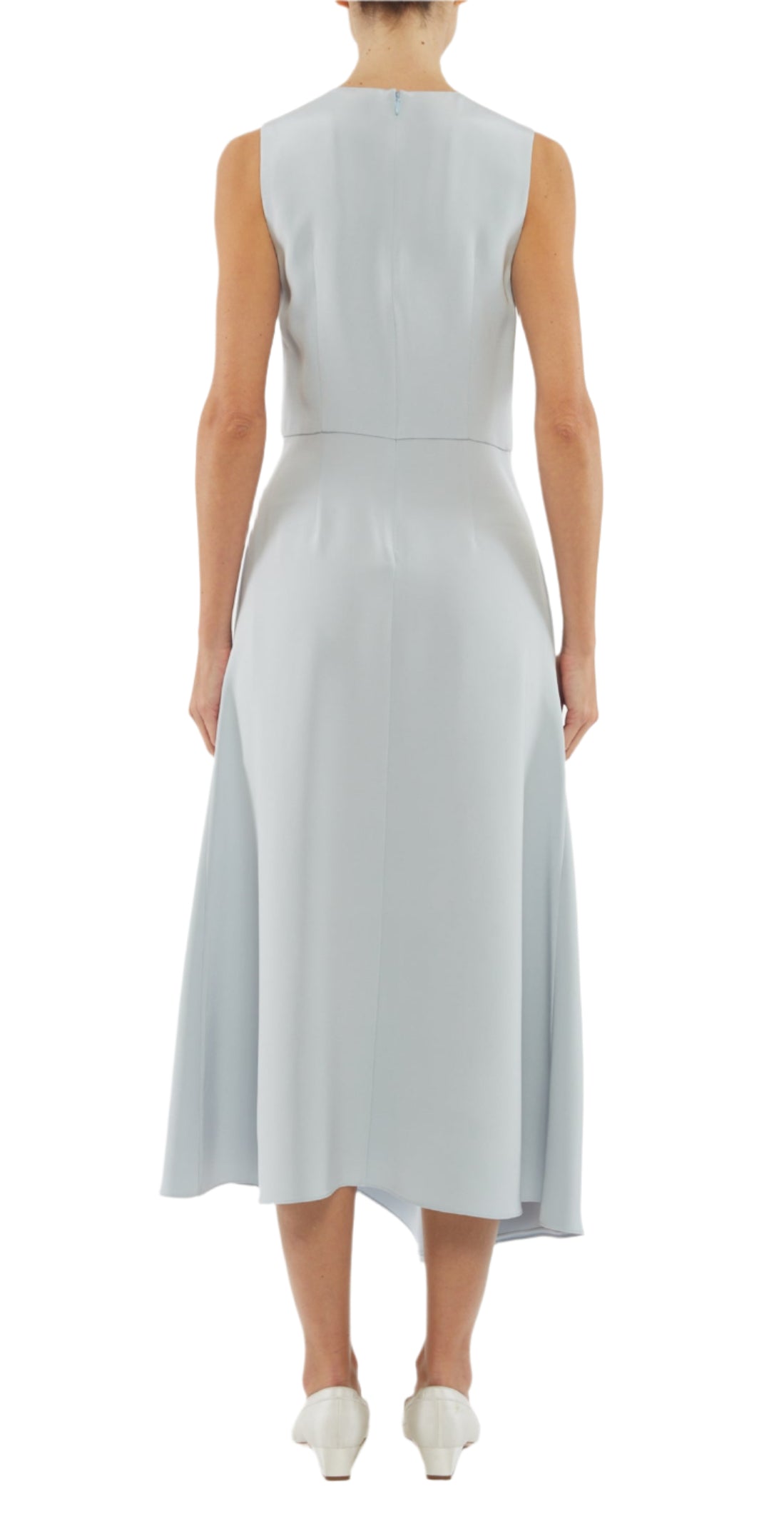 A person wearing the Adam Lippes Lynn Silk Crepe Dress, a sleeveless light blue midi with an asymmetric draped panel, and white shoes stands with their back to the camera against a white background.