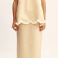 A woman wears the POSSE Essie Column Skirt, a beige premium linen two-piece with scalloped white trim, paired with white open-toe sandals.