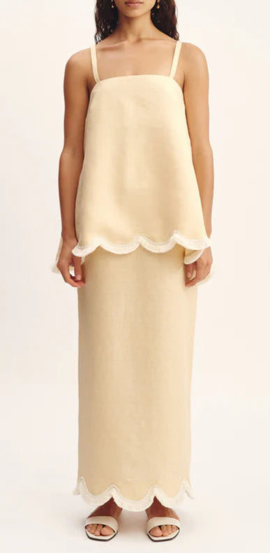 A woman wears the POSSE Essie Column Skirt, a beige premium linen two-piece with scalloped white trim, paired with white open-toe sandals.