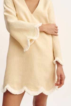 Wearing the Essie Kaftan by POSSE, this style features a light yellow linen fabric, long sleeves, deep v-neckline, and scalloped hems with white fringe trim—ideal for a chic vacation dress look.