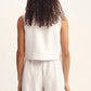 A person with curly hair is shown from behind, wearing POSSE's Rio Top—a sleeveless white high-neck top—paired with matching relaxed-fit linen pants, set against a plain background.
