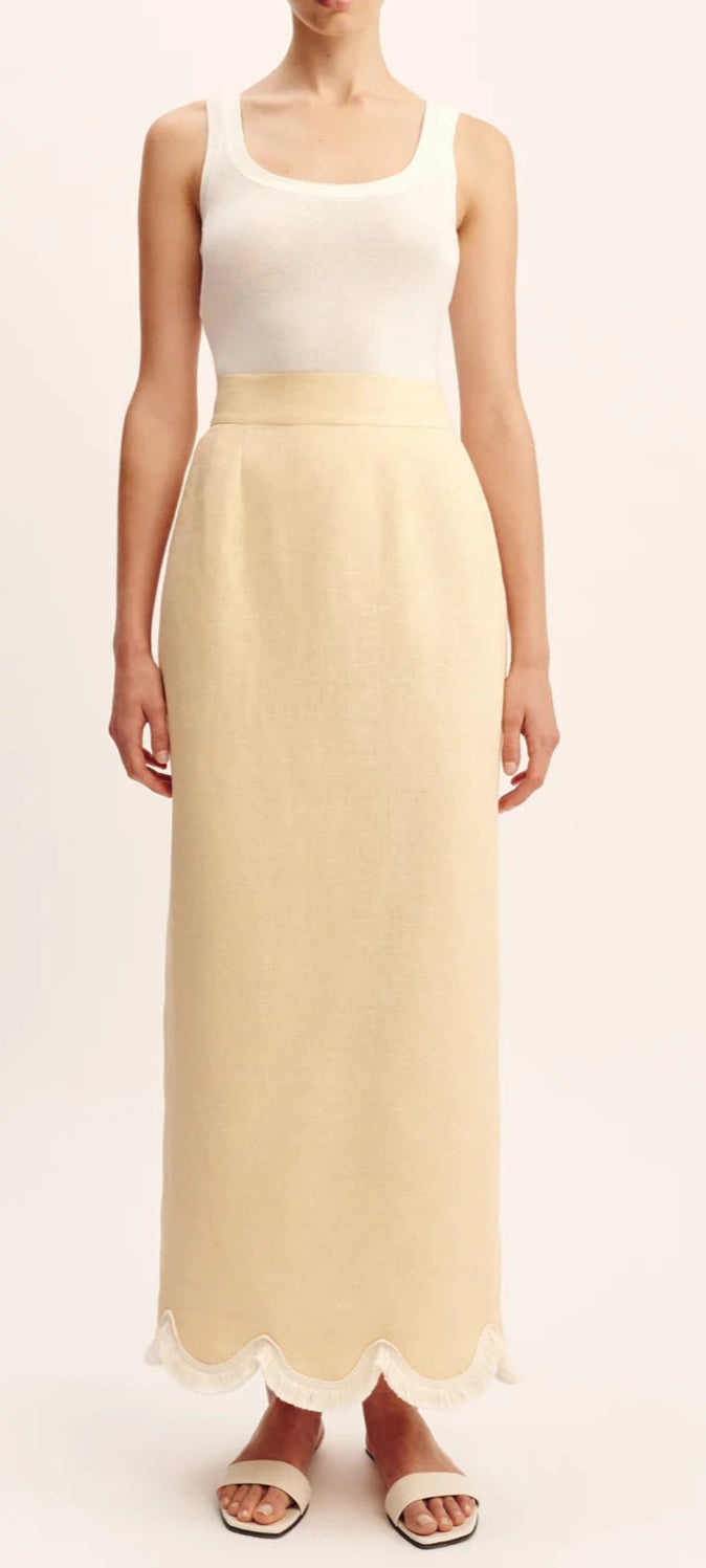 A person wears a sleeveless white top, the POSSE Essie Column Skirt in cream—with a high-rise waist and scalloped hem—and white sandals.