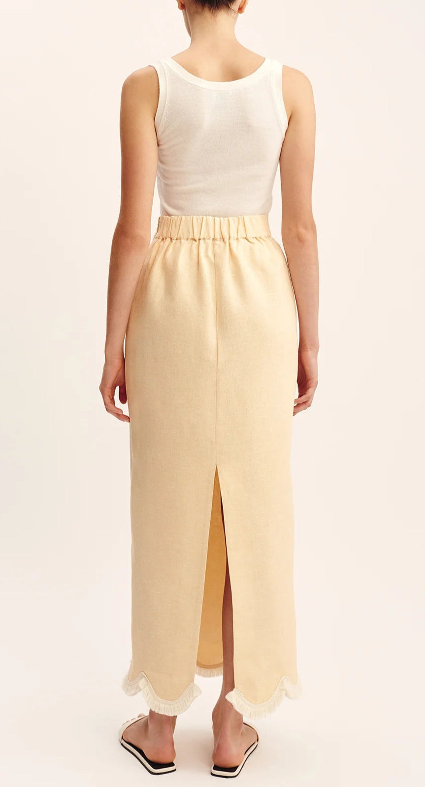 A person stands in flat sandals, photographed from behind, wearing a white tank top and the POSSE Essie Column Skirt featuring a high-rise waist, back slit, and fringe hem.