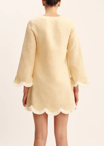 A person is seen from the back wearing the POSSE Essie Kaftan, a light yellow linen dress featuring long sleeves, scalloped edges, and lace trim at the cuffs and hem.
