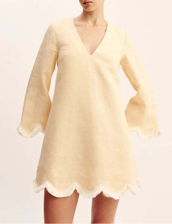 A woman wears the POSSE Essie Kaftan, a light beige long-sleeve vacation dress with a deep V-neckline and scalloped hem and sleeve edges trimmed with white fringe.