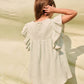 A young girl stands outdoors with her back to the camera, wearing the Louise Misha Girls’ Taliana Dress—a light green cotton gauze dress featuring ruffled sleeves—while holding her hair with one hand.
