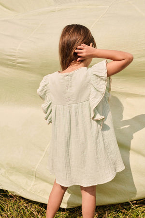 A young girl stands outdoors with her back to the camera, wearing the Louise Misha Girls’ Taliana Dress—a light green cotton gauze dress featuring ruffled sleeves—while holding her hair with one hand.