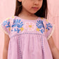 A young girl wearing the Louise Misha Girls’ Talula Dress—featuring light purple fabric and blue, yellow, and pink textured embroidery—stands against a pink background, looking down.