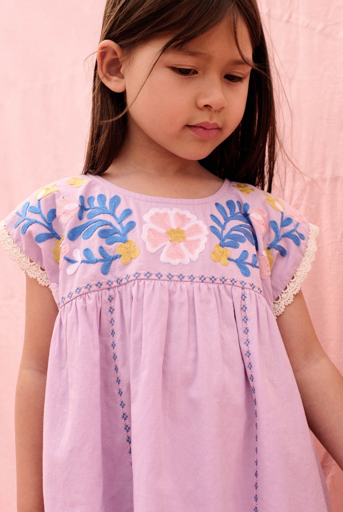 A young girl wearing the Louise Misha Girls’ Talula Dress—featuring light purple fabric and blue, yellow, and pink textured embroidery—stands against a pink background, looking down.