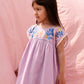 A young girl wearing the Louise Misha Girls’ Talula Dress in light purple with textured embroidery stands in front of a pink fabric backdrop, gazing to the side.