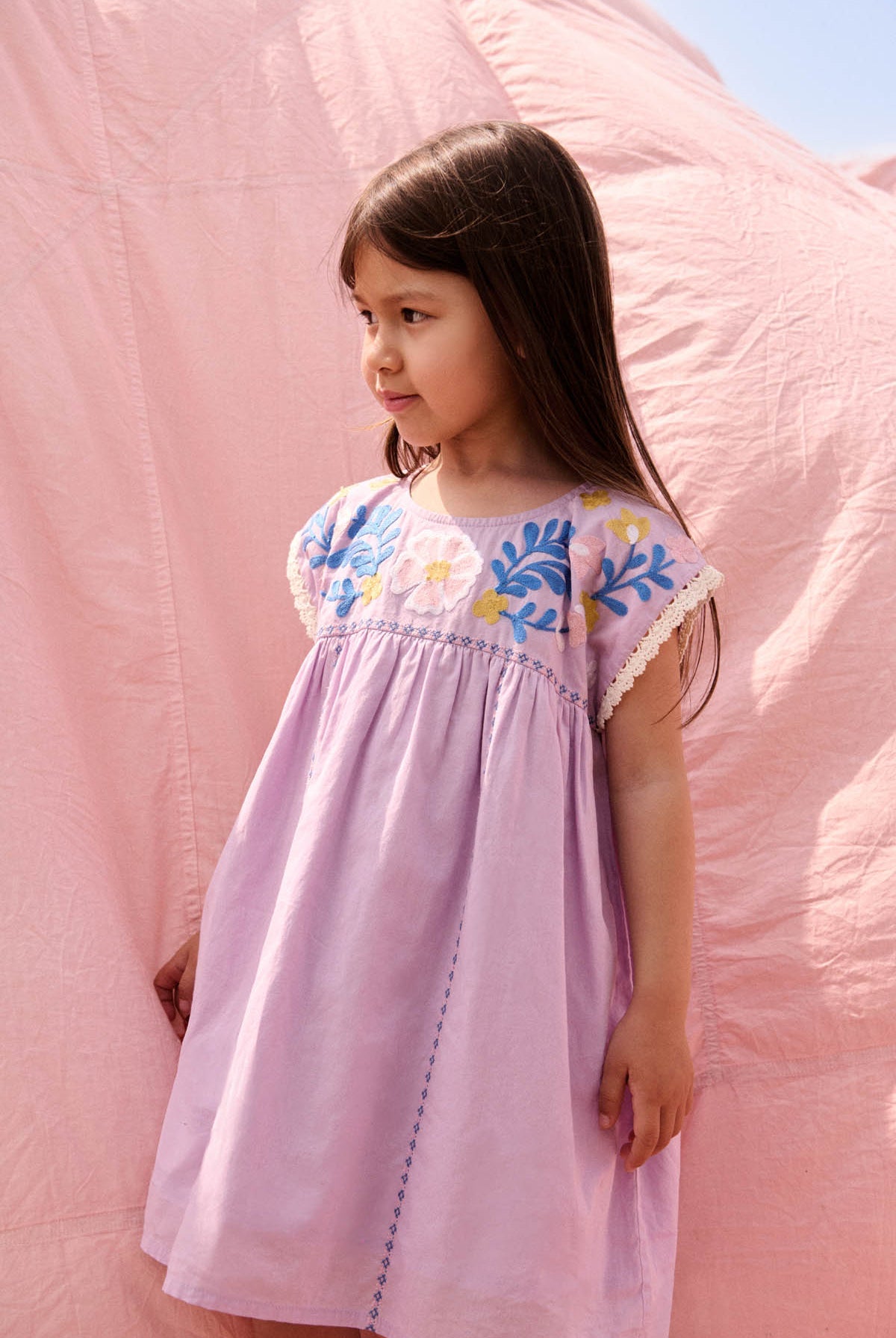 A young girl wearing the Louise Misha Girls’ Talula Dress in light purple with textured embroidery stands in front of a pink fabric backdrop, gazing to the side.