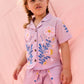 A young child with braided hair wears the Louise Misha Girls’ Alova Shirt—a light pink, short-sleeved floral top—along with matching shorts, standing in front of a pale pink fabric backdrop.