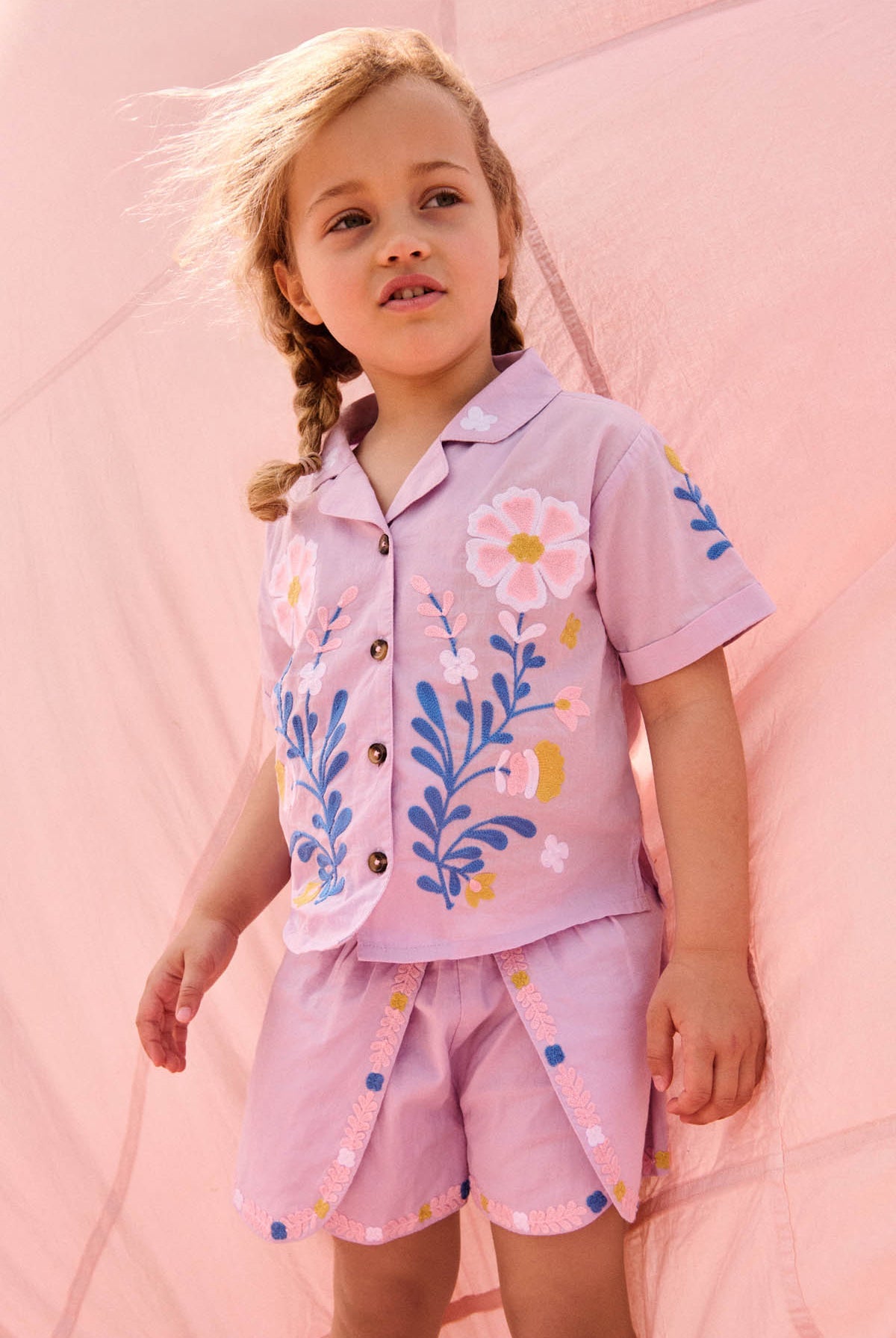A young child with braided hair wears the Louise Misha Girls’ Alova Shirt—a light pink, short-sleeved floral top—along with matching shorts, standing in front of a pale pink fabric backdrop.